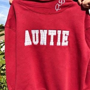 May include: A red mock neck sweatshirt featuring the word AUNTIE in white-bordered, beige and white checkered block letters across the chest. The collar area includes white embroidered script lettering of the initials QB on the right side.