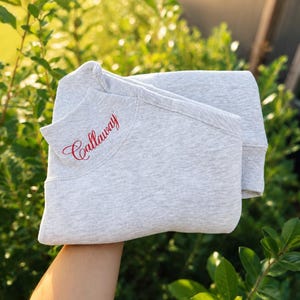 May include: A light gray heathered sweatshirt featuring the word Callaway embroidered in red script on the upper left chest area. The garment has a ribbed mock neck collar and ribbed cuffs, showcasing a clean and simple design.