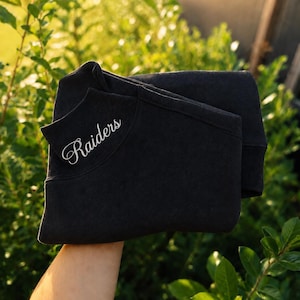 May include: A charcoal black sweatshirt featuring the word Raiders embroidered in a white cursive script on the upper left chest area. The garment has a ribbed crew neckline and long sleeves, with a folded construction visible in the center.