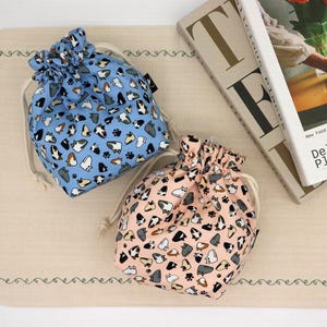 May include: Two drawstring pouches made of fabric, one in light blue and one in soft pink. Both bags feature a repeating pattern of small, stylized cat faces in various colors and black paw prints, finished with cream-colored cord drawstrings.