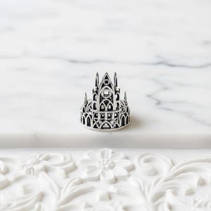 Può includere: Un anello in argento caratterizzato da un intricato design traforato che imita la struttura architettonica di una cattedrale gotica, completo di archi a sesto acuto, guglie alte e dettagliati motivi a traforo su tutta la fascia.