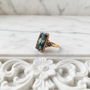May include: A gold ring featuring a rectangular labradorite gemstone with a blue iridescent flash. A small, diamond-shaped accent is set in the center of the stone. The gold band has decorative metalwork details on the sides of the setting.