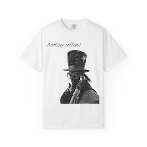 May include: A white short-sleeve t-shirt featuring a black and white graphic print of Bootsy Collins wearing a top hat and star-shaped glasses. The text "bootsy collins" is printed in a black cursive font above the graphic on the chest area.