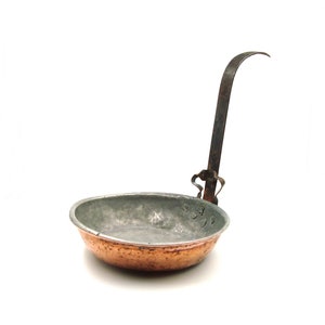 May include: A small, antique copper pan with a long, curved iron handle. The pan has a rounded bowl shape with a silver-colored interior and a copper exterior. The handle is attached to the pan with decorative metalwork. The pan is likely for cooking.