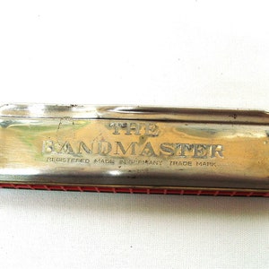 以下が含まれることがあります： 銀色の金属カバーと赤い櫛が付いたヴィンテージハーモニカ。「THE BANDMASTER」の文字がカバーに刻印されており、「REGISTERED MADE IN GERMANY TRADE MARK」も記載されています。ハーモニカは長方形です。