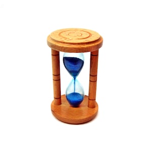 May include: A wooden hourglass with a light brown finish. The hourglass has a clear glass center filled with blue sand. The hourglass is supported by four wooden pillars and has a circular top and bottom.