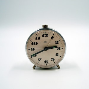 May include: A vintage silver-toned alarm clock with a cream-colored face and black numerals. The clock has a small, round design with a metal casing and three small feet. The hands are black with a green accent. The text "Made in Czechoslovakia" is visible.