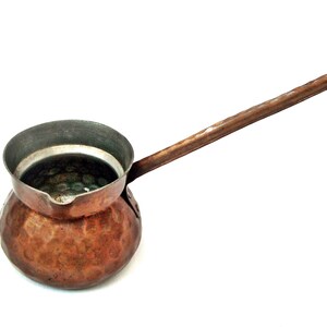 Puede incluir: Una cafetera de cobre con un mango largo. La olla tiene una base redondeada y una parte superior ancha y abierta. El mango está hecho del mismo material de cobre y se extiende desde el lado de la olla. La olla está diseñada para preparar café.
