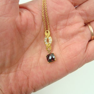 May include: A gold-colored necklace with a pendant featuring a square, dark red gemstone. The pendant is suspended from a gold chain with a decorative infinity-shaped design adorned with small, clear gemstones.