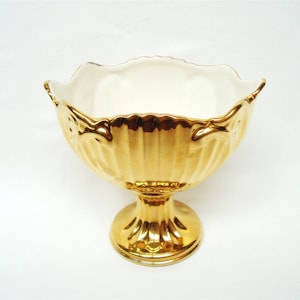 Könnte beinhalten: Eine goldfarbene Schale mit Fuß und gewelltem Rand. Die Schale hat ein geriffeltes Design auf der Außenseite und einen weißen Innenraum. Die Schale ist wahrscheinlich aus Keramik oder Porzellan und eignet sich zum Servieren von Obst oder zu dekorativen Zwecken.
