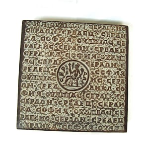 May include: A square, brown ceramic tile featuring white text and a central circular emblem. The text is in a foreign language, and the emblem shows a figure with a bow and arrow. The tile has a rustic, aged appearance.