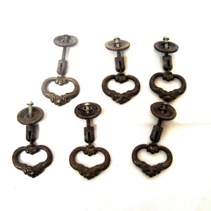 May include: A set of six antique-style bronze-toned metal drawer pulls. Each pull features a circular mounting plate with a central screw and a hinged, heart-shaped drop handle with ornate, raised detailing along the bottom edge.