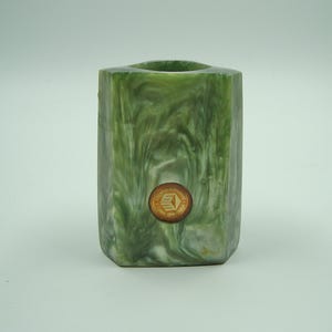 Puede incluir: Un portalápices de resina verde y blanca veteada con forma hexagonal. Una pequeña pegatina redonda de color dorado está adherida a la parte delantera. El portalápices tiene un interior hueco.