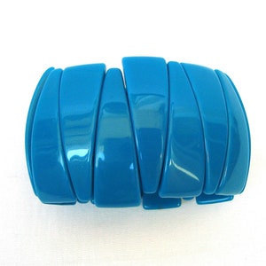 May include: A vibrant turquoise bracelet composed of segmented, curved pieces. The glossy, articulated design allows for flexibility and a comfortable fit. The bracelet's bold color and unique construction make it a striking accessory.