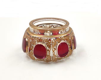 May include: A clear glass jar with a gold rim and ornate gold filigree design. The jar features five red circular accents. The jar is likely a decorative item or a container for small items.
