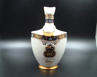 Puede incluir: Una botella de cerámica blanca con detalles dorados y patrones florales azules. La botella tiene una forma única y presenta el texto "Year of the Dog" en letras doradas. Una etiqueta dorada cuelga del cuello.