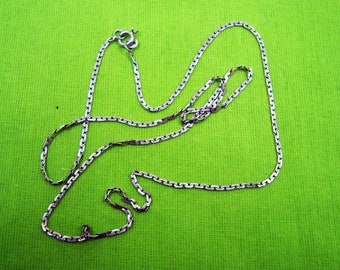 May include: A silver-colored necklace with a delicate chain design, featuring a clasp closure. The necklace is displayed against a bright green background, highlighting its metallic sheen. The chain is composed of small, interconnected links.