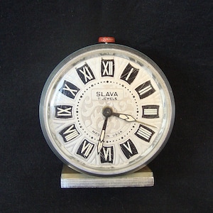 May include: A vintage Slava alarm clock with a silver face and black Roman numerals. The clock has a red top button and a silver metal base. The face reads "SLAVA 11 JEWELS" and "MADE IN USSR".