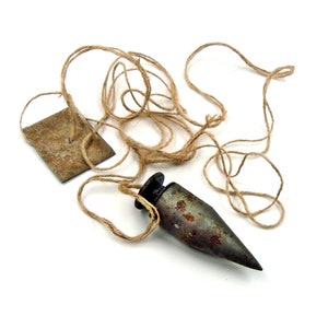 May include: A vintage plumb bob with a pointed end, attached to a length of twine, and a small, weathered tag. The metal bob has a rusted appearance, suggesting age and use. The twine is a natural, light brown color.