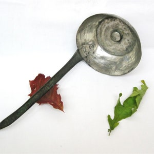 May include: A vintage metal ladle with a long, curved handle and a circular bowl. The ladle is a dull silver colour, with a dark handle. A red leaf and a green leaf are next to the ladle.
