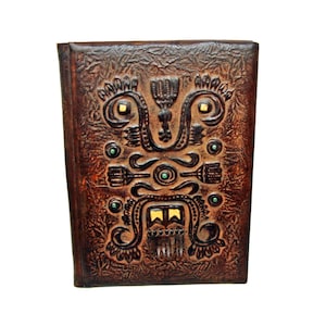 May include: A brown leather journal with an embossed Aztec-inspired design. The cover features a central symmetrical pattern with gold and green accents. The journal has a textured surface and a dark brown spine.