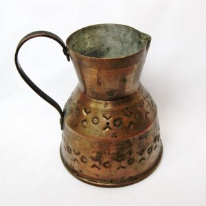 May include: A vintage copper pitcher with a curved handle. The pitcher has a flared top and a decorative pattern of circles and crosses around the base. The copper has a warm, aged patina, suggesting it is an antique or a well-loved piece.
