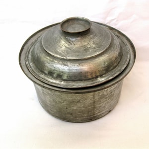 May include: A vintage metal cooking pot with a lid. The pot is a dull silver color with a textured surface. The lid has a small handle on top and fits snugly over the pot. The pot appears to be made of a heavy metal.