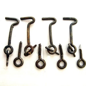 Puede incluir: Un conjunto de cuatro ganchos de puerta y tornillos de ojo de metal antiguos. Los ganchos tienen una parte superior curvada y un lazo en la parte inferior. Los tornillos de ojo tienen un ojal circular. El metal tiene un aspecto rústico y envejecido.