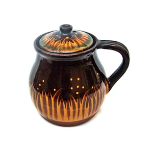 Può includere: Un vaso di ceramica marrone con coperchio e manico. Il vaso è decorato con disegni a forma di erba arancione e gialla e piccoli punti gialli. Il coperchio ha un design simile con un pomello marrone scuro. Il vaso viene probabilmente utilizzato per cucinare o servire.