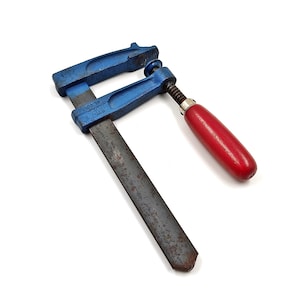 May include: A blue and steel bar clamp with a red wooden handle. The clamp has a threaded screw mechanism for tightening and is designed for woodworking or construction projects. The metal components show signs of wear.
