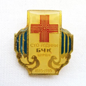 Könnte beinhalten: Vintage Anstecknadel mit goldenem Rand und rotem Kreuz. Die Anstecknadel zeigt Text in einer Fremdsprache mit den Jahren 1885-1985 unten. Zwei Tauben befinden sich am unteren Rand der Anstecknadel, mit blauen Akzenten an den Seiten.