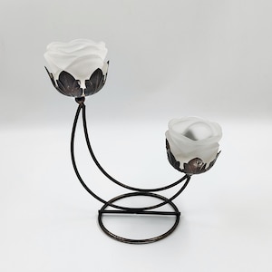 May include: A decorative candle holder with two frosted glass rose-shaped candle cups. The cups are supported by a dark brown, wrought iron-style frame with a curved design. The base is circular.