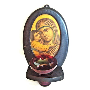 Puede incluir: Un icono religioso que representa a María y Jesús, enmarcado en un marco ovalado de madera oscura. Debajo del marco hay un pequeño cuenco redondo de cristal morado oscuro. La imagen tiene un agujero en la parte superior para colgar.