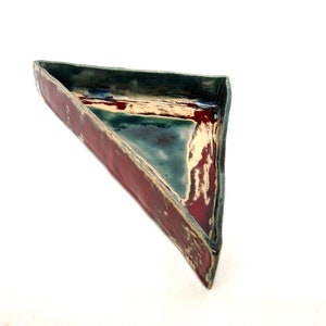 Puede incluir: Un plato triangular de cerámica hecho a mano con exterior rojo e interior turquesa. El borde es verde oscuro con toques azules. Ideal para servir o exhibir.