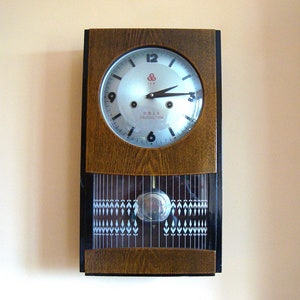 May include: A rectangular wooden wall clock with a silver face and black hands. The clock face has black numbers and the text "SHANGHAI CHINA". A pendulum swings below the clock face.