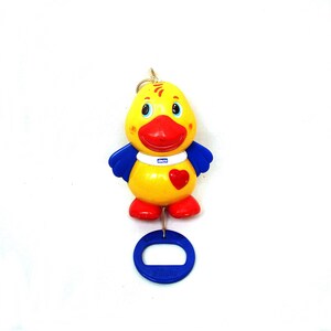 May include: A yellow and blue duck-shaped baby toy with a red beak, feet, and a heart on its chest. The toy has blue wings and a blue ring attached to the bottom. The brand name "chicco" is printed on the toy.