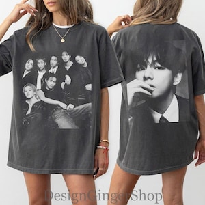 Puede incluir: Una camiseta de manga corta gris carb&oacute;n con un estampado gr&aacute;fico en blanco y negro de un grupo de siete personas en la parte delantera y un gran retrato en blanco y negro de una persona en la parte trasera. Tiene cuello redondo y corte holgado.