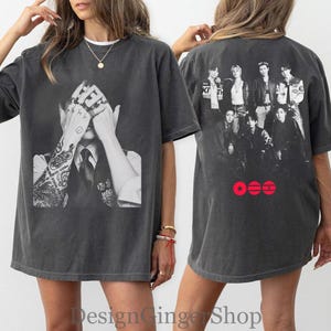 May include: A charcoal gray oversized t-shirt featuring a black and white graphic on the front of a person with tattooed arms covering their face. The back displays a black and white group photograph of seven individuals with three red circular icons below.