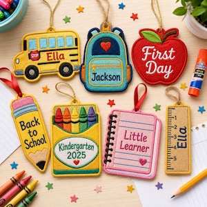 May include: A collection of seven embroidered felt ornaments featuring school-themed designs including a yellow bus with the name Ella, a blue backpack with the name Jackson, a red apple with First Day, a pencil, a crayon box, a notebook, and a ruler.