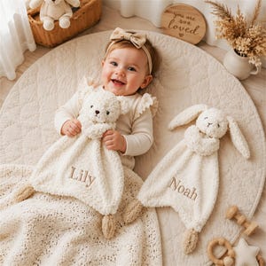May include: Two cream-colored plush security blankets featuring embroidered names. One blanket is shaped like a teddy bear with the name Lily, and the other is shaped like a rabbit with the name Noah. Both are made of soft, textured fabric.