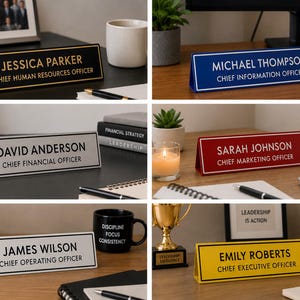 May include: A collection of six rectangular desk nameplates in black, blue, white, red, and yellow. Each nameplate features a person's name and job title in a contrasting font color, with a border matching the text color on the face of the sign.