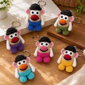 May include: Six crocheted keychains shaped like potato figures, each featuring a black hat, white arms, pink ears, and large white eyes with black pupils. The figures have noses in red or orange and wear trousers in blue, green, purple, pink, or yellow.
