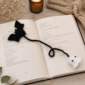 Può includere: Un segnalibro all'uncinetto fatto a mano con un pipistrello nero a un'estremità e un piccolo fantasma bianco con occhi neri all'altra, collegati da un lungo cordoncino nero all'uncinetto sulle pagine aperte di un libro con testo stampato.