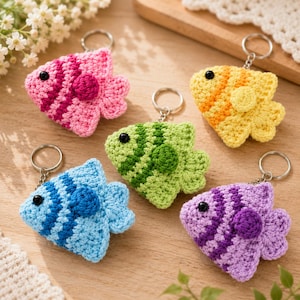 May include: Five crocheted fish keychains in pink, yellow, green, blue, and purple. Each fish features a striped pattern, a small round side fin, and a black bead eye. A silver metal split ring is attached to the top of each fish for hanging.
