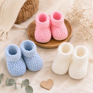 Könnte beinhalten: Drei Paar handgehäkelte Baby-Schühchen in einfarbigen Pastelltönen. Ein Paar ist hellrosa, ein Paar hellblau und ein Paar cremeweiß. Jedes Paar verfügt über eine abgerundete Zehenpartie, ein dickes geripptes Bündchen und eine strukturierte Garnoptik.