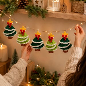 May include: A festive garland featuring five crocheted Christmas tree ornaments strung on a thin wire with small LED lights. Each tree is green with white trim, a yellow star topper, and a small red bow. The trees alternate between dark and light green.