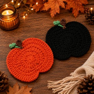 May include: Two round crochet coasters shaped like pumpkins, one in bright orange and one in solid black. Each coaster features a small brown stem and two tiny green leaves attached to the top edge, crafted with a textured yarn stitch pattern.