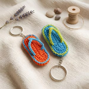 May include: Two crochet flip-flop keychains. One keychain features an orange sole with light blue straps, while the other has a blue sole with lime green straps. Each keychain includes a silver-toned metal split ring attached by a short chain.