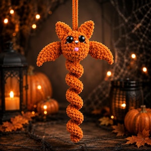 May include: A hanging orange crochet bat ornament featuring a textured spiral tail, large black plastic eyes, and a small pink stitched mouth. The plush creature is crafted from soft yarn and suspended by a matching orange cord for display.
