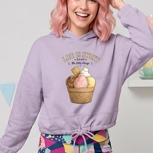 Op de afbeelding: Een lichtpaarse hoodie met lange mouwen, een trekkoord in de zoom en een afbeelding van een ijshoorntje met de tekst "LIFE IS SWEET ENJOY the little things". De hoodie wordt gecombineerd met een legging met een veelkleurig geometrisch en bloemenpatroon.